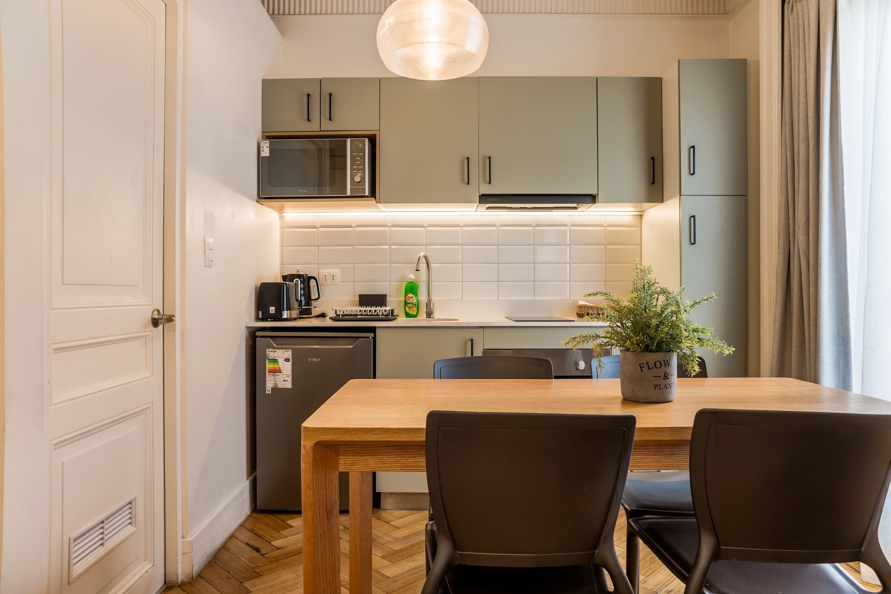 Een kleine keuken met een magnetron, spoelbak en kastjes, een eettafel met stoelen, en een potplant.