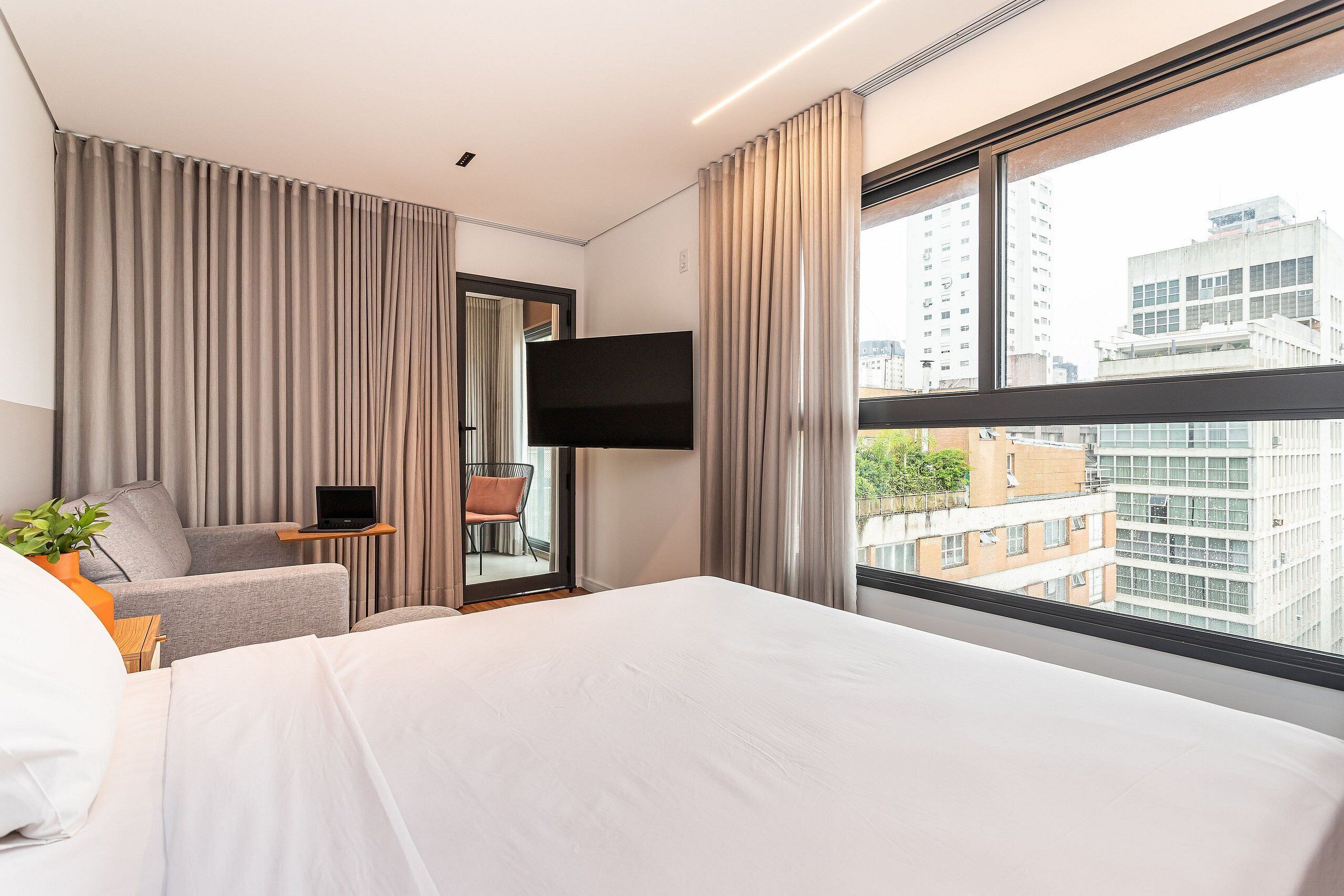 Habitación de hotel con cama, escritorio con computadora portátil, silla, televisor montado en la pared y ventana grande con vista a la ciudad.