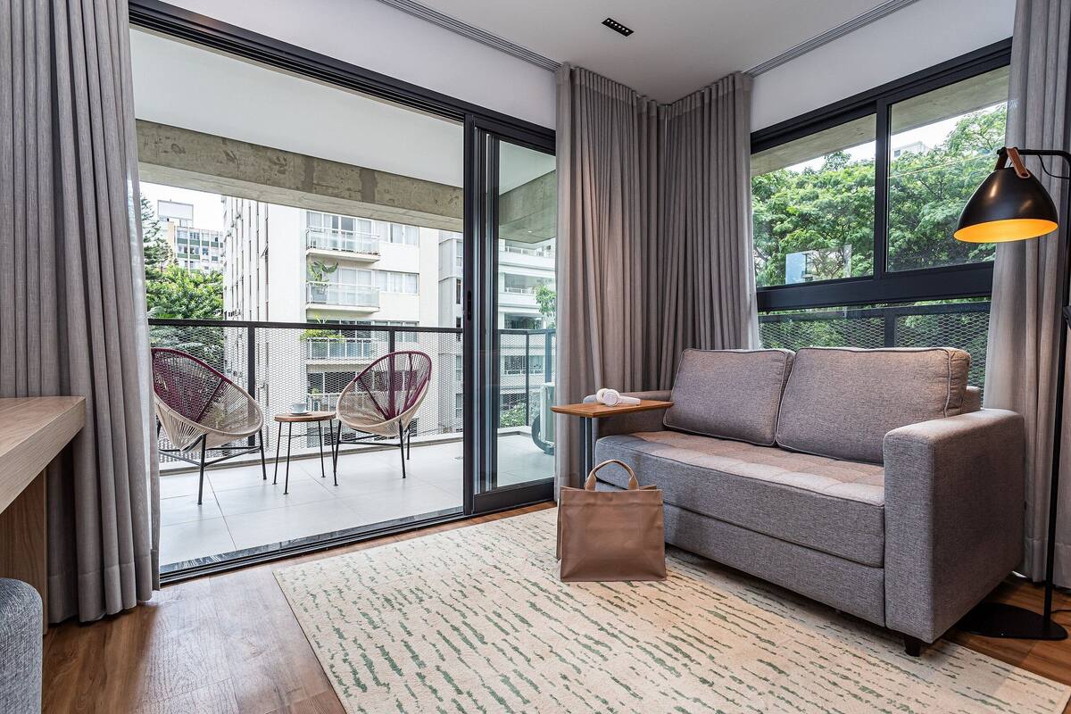 A modern living room with a sofa, a coffee table, a chair, a large window, and a balcony with a view of buildings and trees.