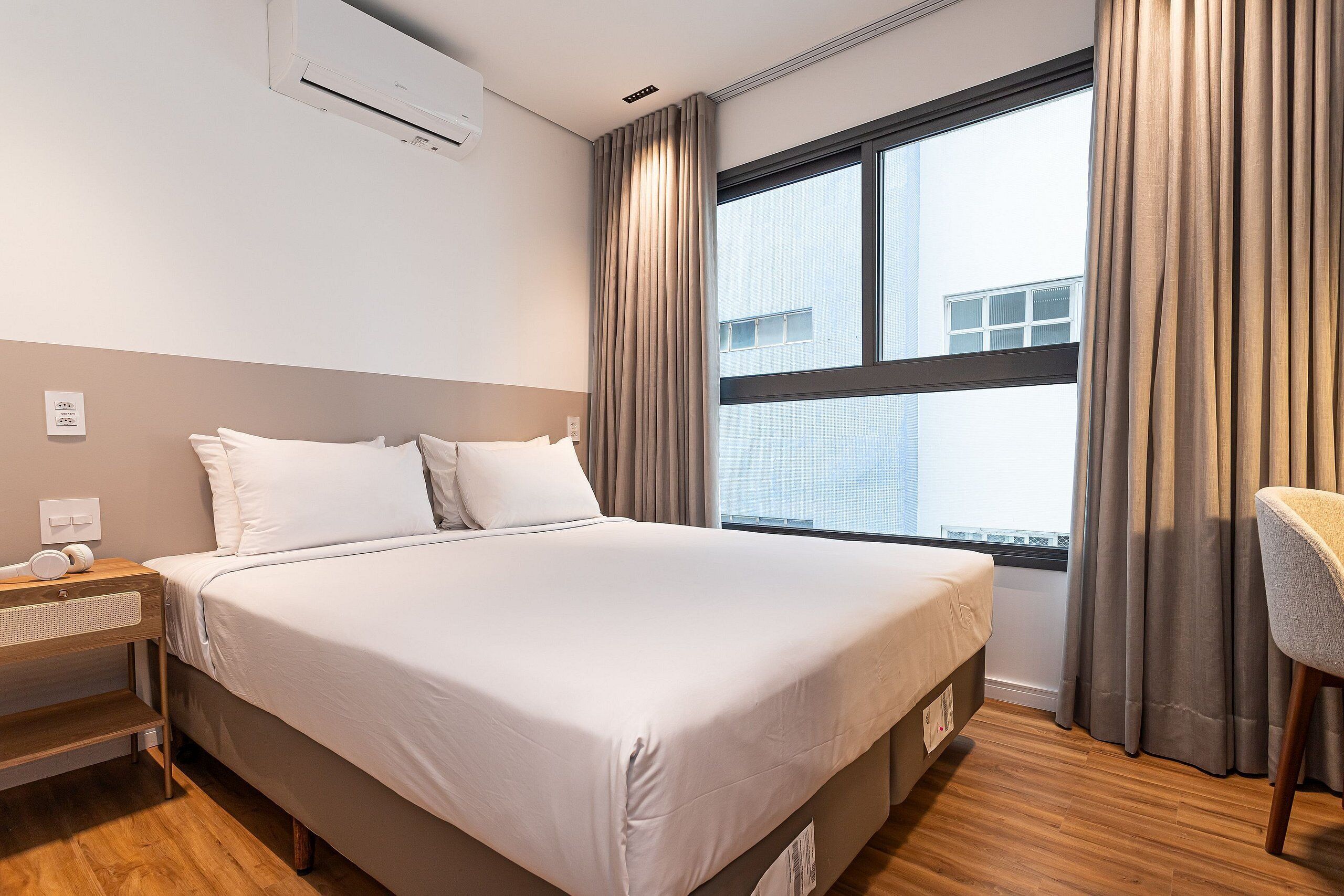 A hotel room with a large bed, a bedside table with a lamp, a chair, and a window with curtains.