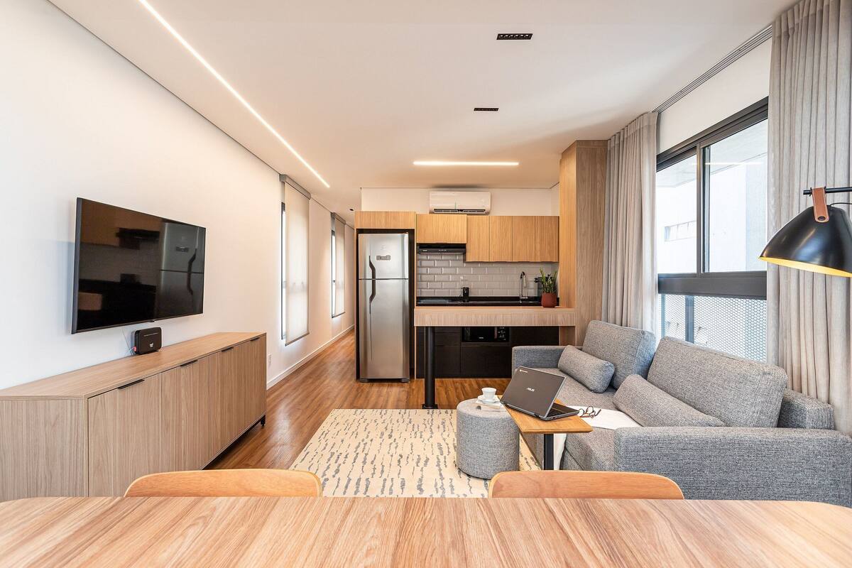 A modern living room with a flat-screen TV, a grey sofa, a wooden dining table, and a kitchen in the background.