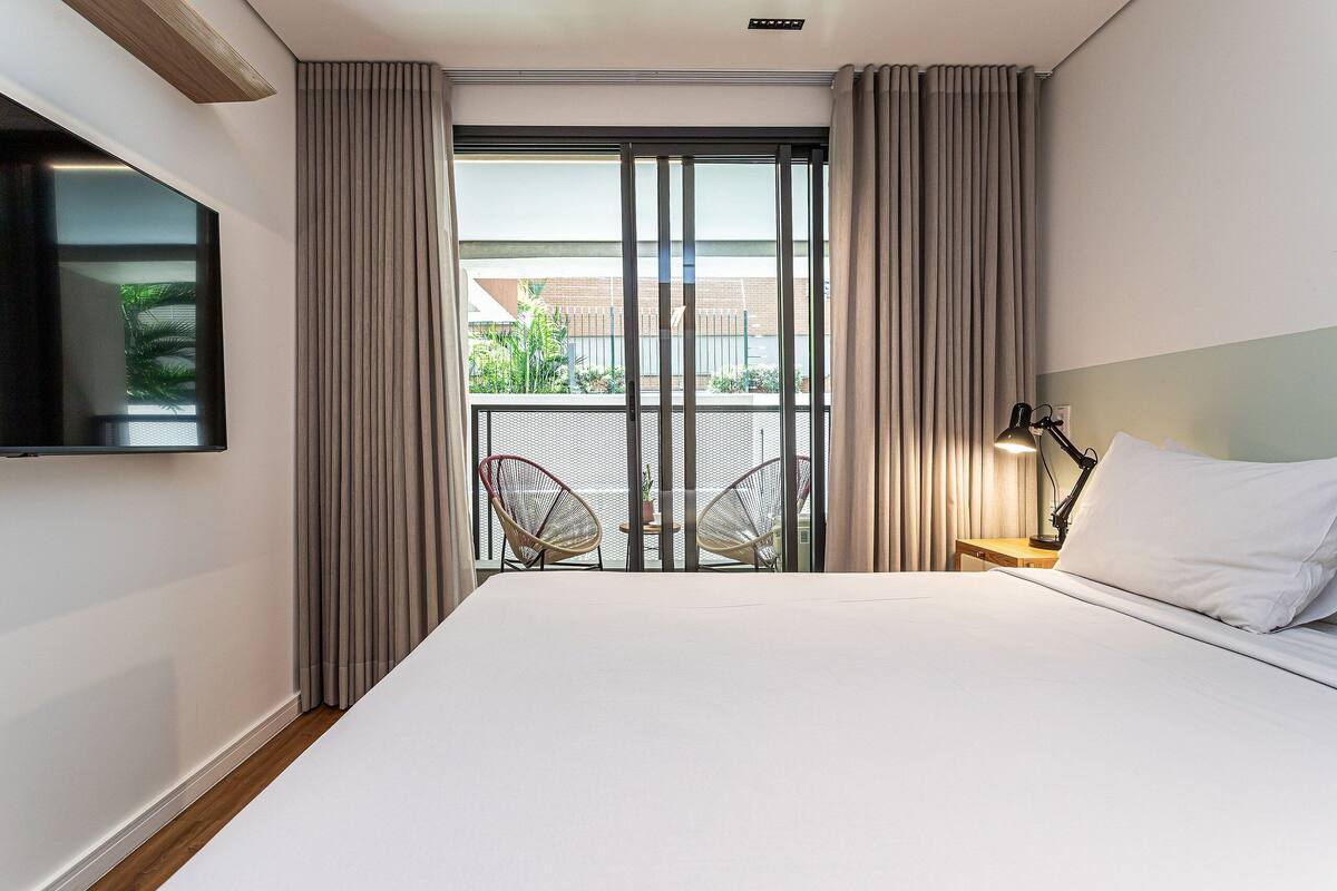 A hotel room with a bed, a TV mounted on the wall, a nightstand with a lamp, and a balcony with curtains.