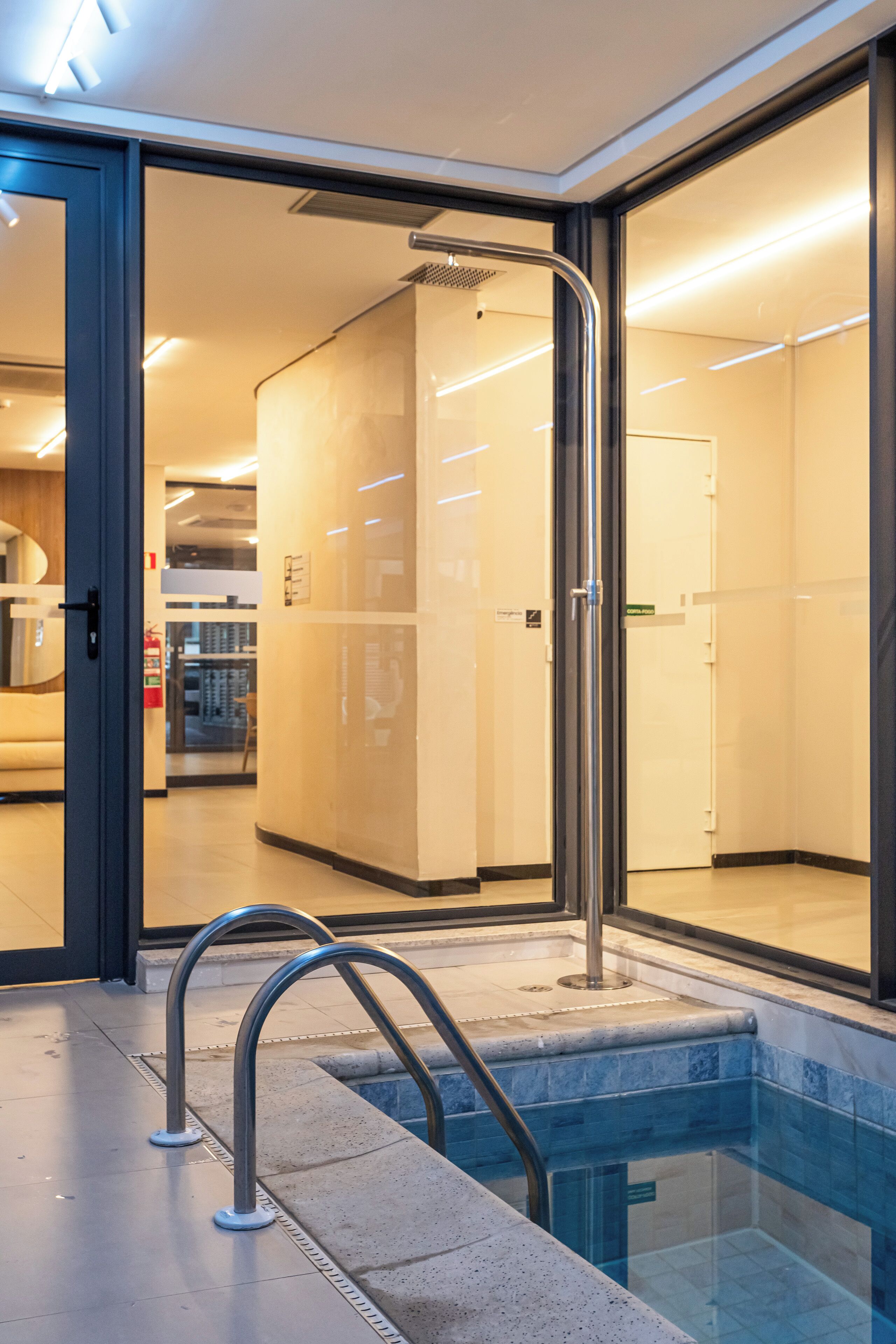 A modern hotel lobby with a glass door leading to a pool area, a visible fire extinguisher, and a clear view of the adjacent restaurant.