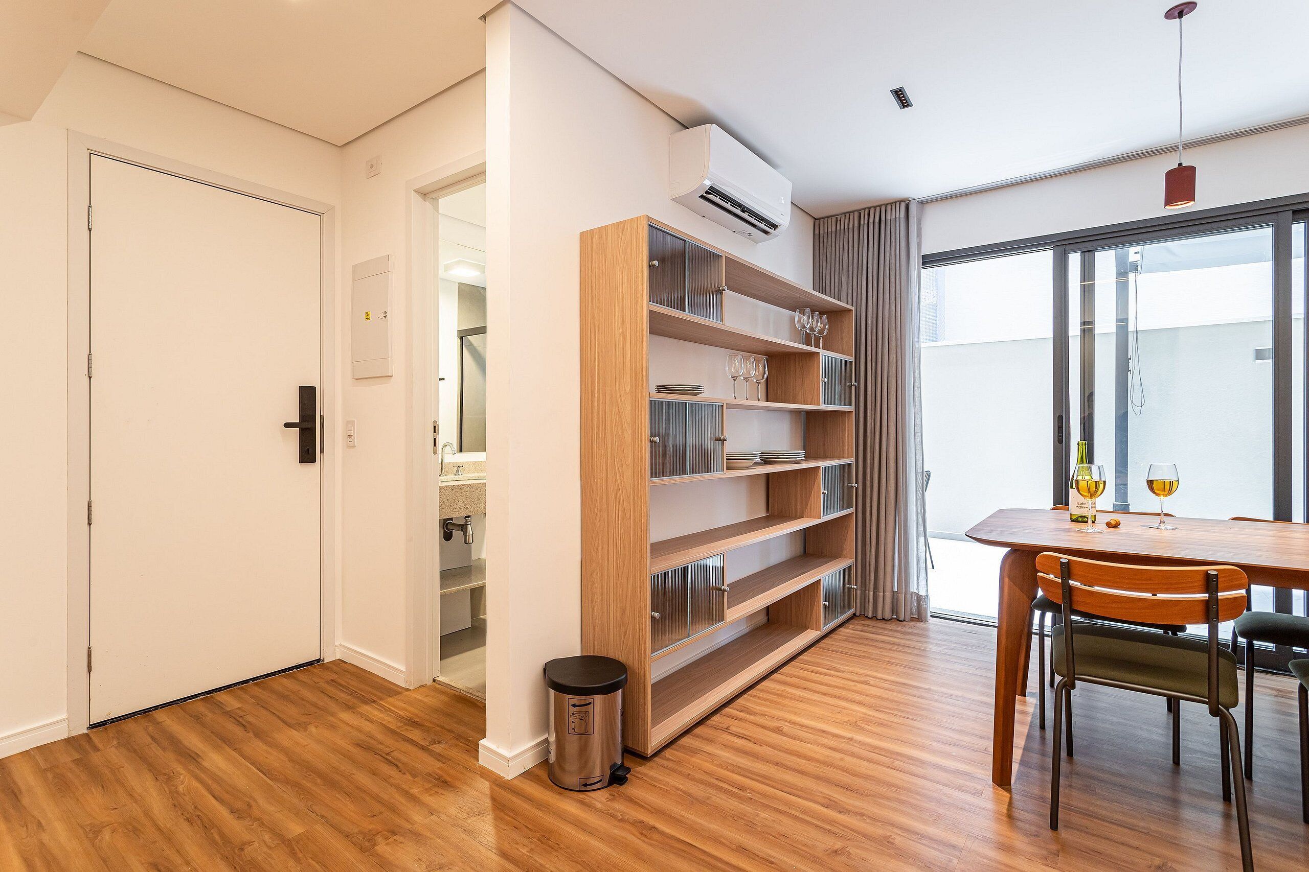 Une pièce moderne avec un plancher en bois, une table à manger avec des chaises, une bibliothèque intégrée et une porte-fenêtre coulissante donnant sur l’extérieur.