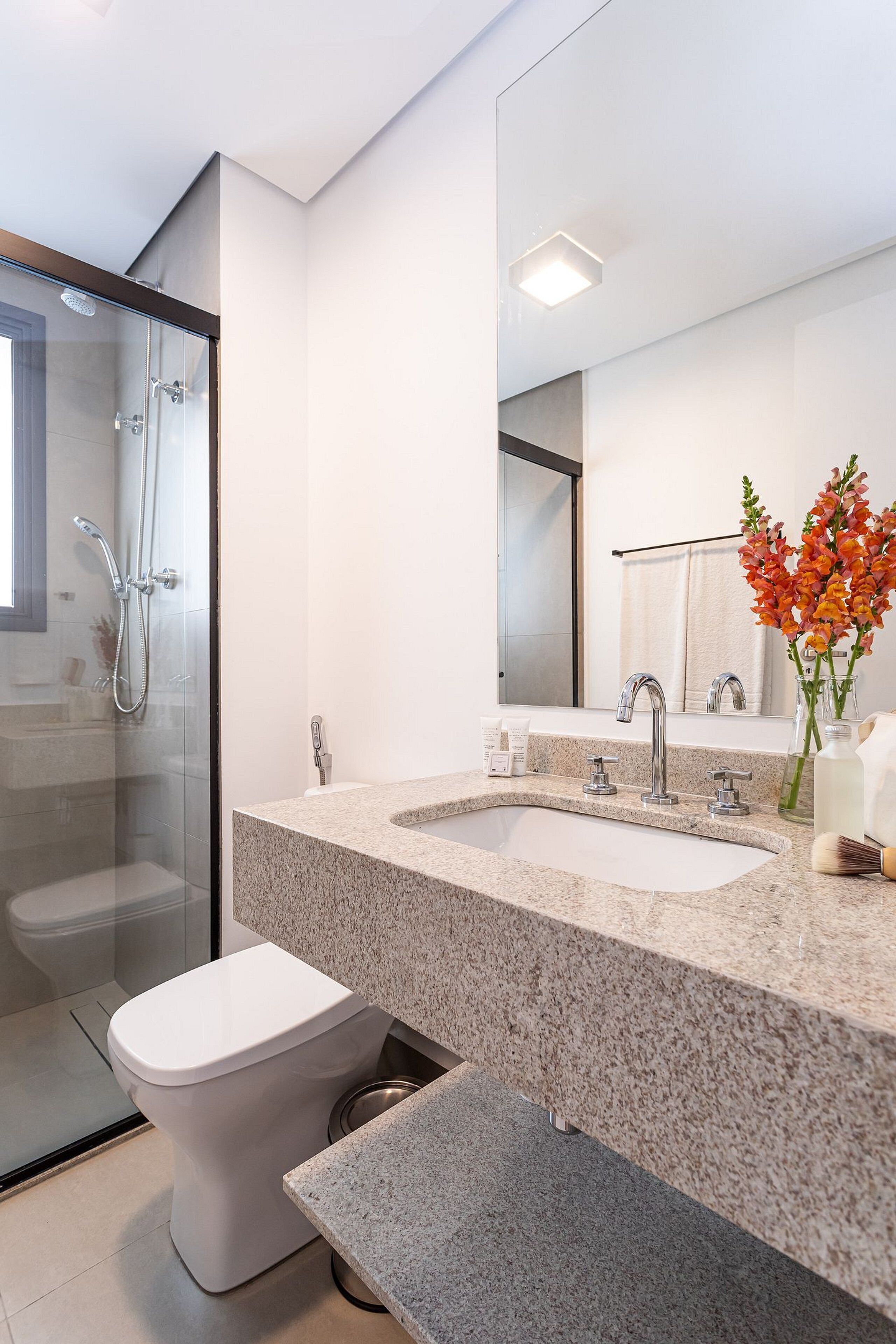 A modern bathroom with a glass shower enclosure, a white toilet, a granite countertop sink, and a mirror reflecting a towel rack.