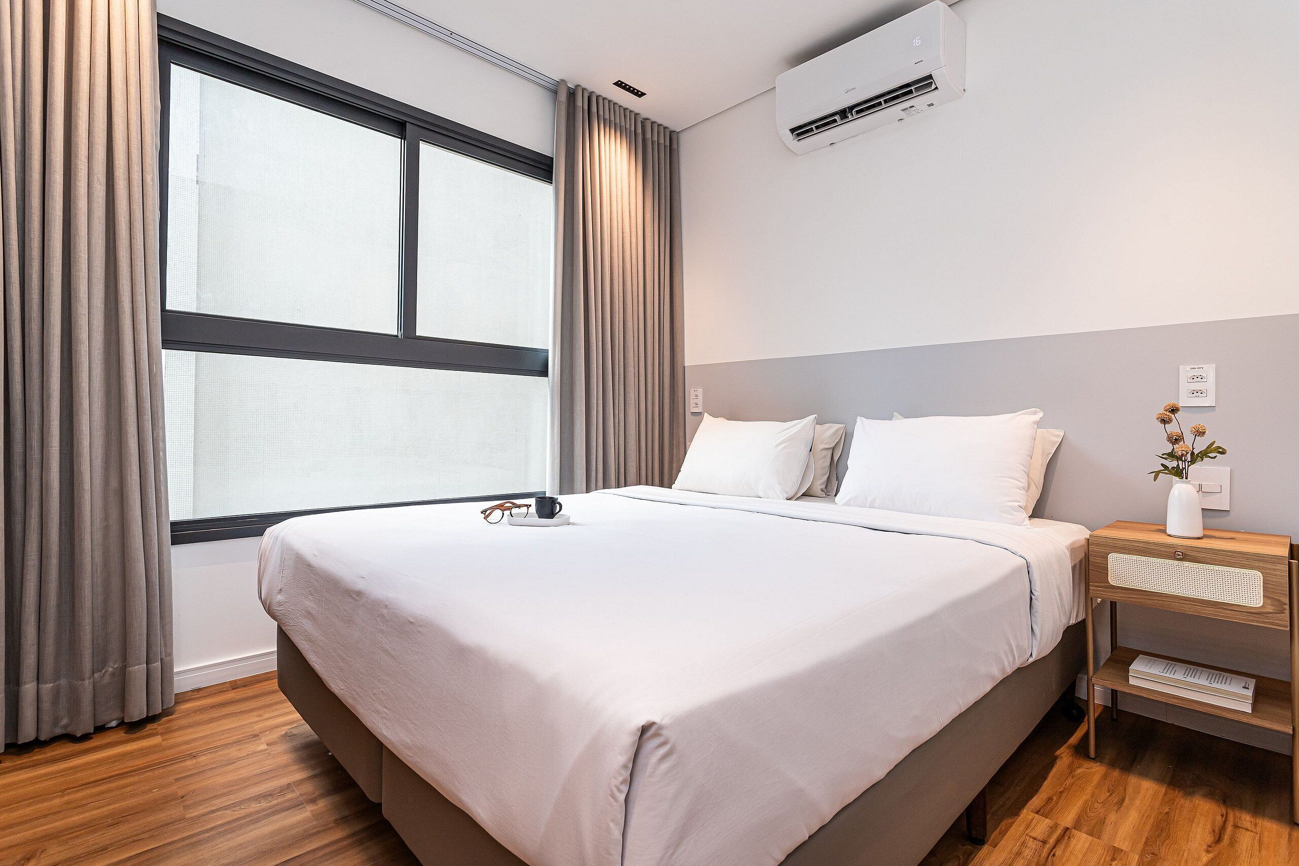 A hotel room with a large bed, a nightstand with a vase, and a window with curtains.