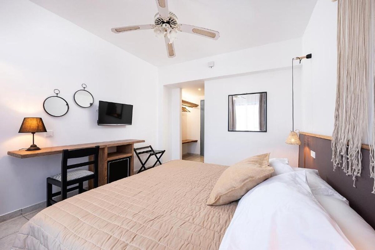 A bedroom with a bed, a desk, a chair, a ceiling fan, a TV, and a framed picture on the wall.