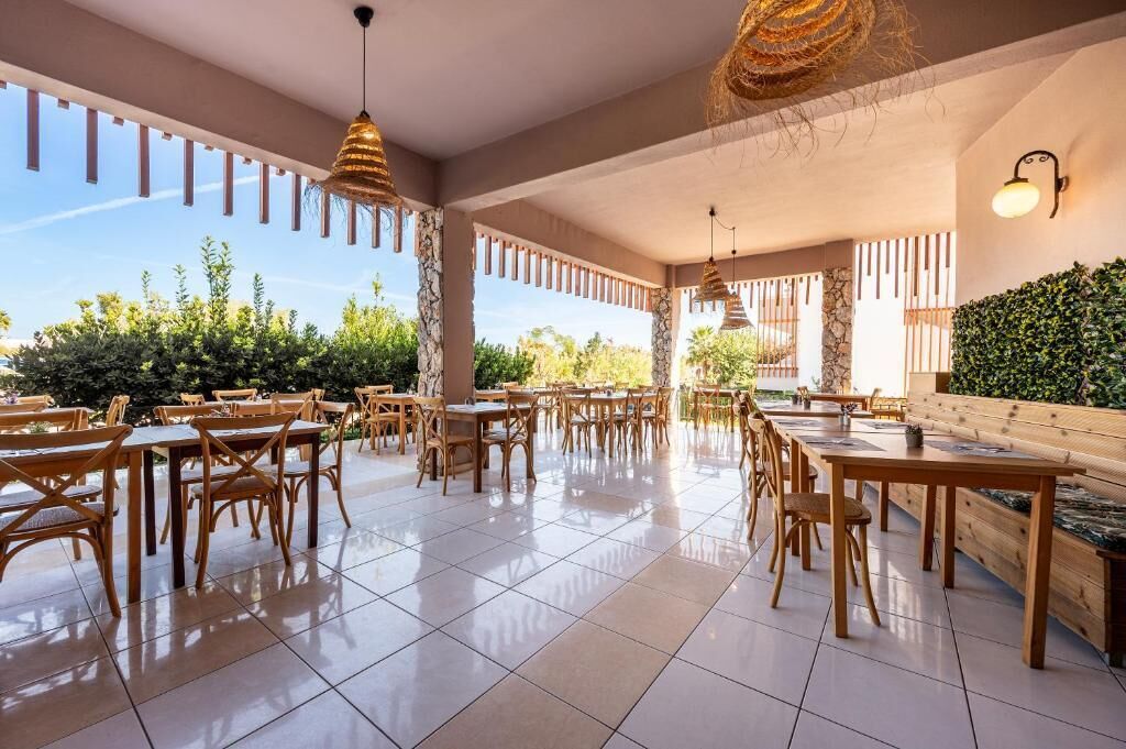 An outdoor dining area with wooden tables and chairs, a pergola with hanging lights, and a view of greenery and trees.