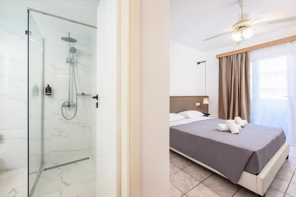 A hotel room with a glass-enclosed shower, a bed with a grey bedspread, and a ceiling fan.
