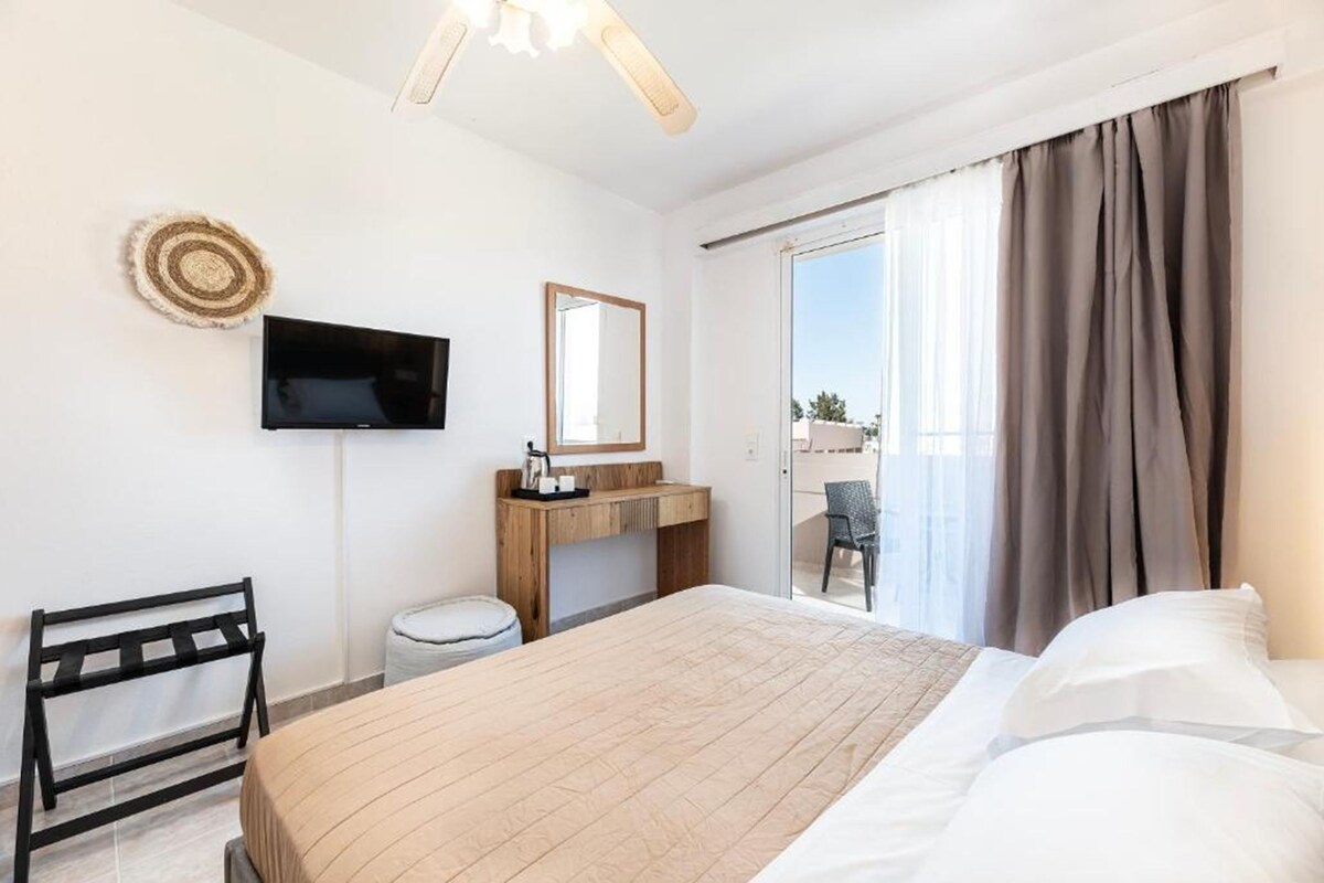 A hotel room with a bed, a TV mounted on the wall, a wooden desk, a chair, and a view of the outdoors through a sliding door.