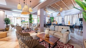 A spacious living area with wicker furniture, a wooden coffee table, and hanging wicker lamps. There is a dining area with blue chairs and a table, and a kitchen in the background. - Aegean Bay Hotel - All Inclusive (Marmari)