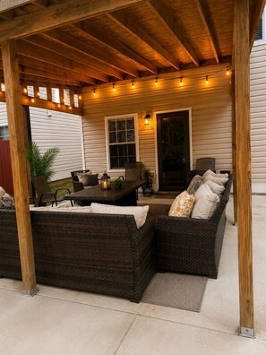 An outdoor patio with a wooden pergola, a wicker sofa, a dining table, and string lights.