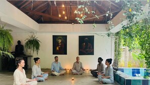 Een groep mensen in een meditatieve houding in een kamer met een hoge plafond, houten balken en hangende planten.