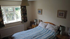 Une chambre à coucher comprenant un lit, deux tables de chevet, une fenêtre avec des rideaux, un radiateur et deux tableaux encadrés accrochés au mur.