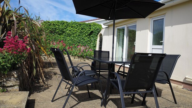 Une terrasse extérieure avec une table et des chaises, un parasol noir, et une maison bordée d’une haie verte et de fleurs roses.