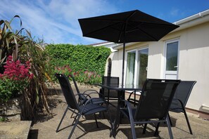 An outdoor patio with a table and chairs, a black umbrella, and a house with a green hedge and pink flowers. - Badgers Cliff (Wadebridge)