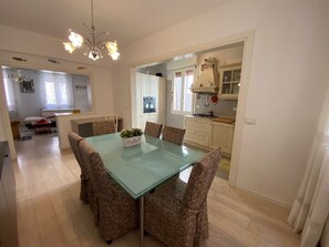Un comedor con una mesa de vidrio y sillas estampadas, una cocina con armarios blancos y una araña.