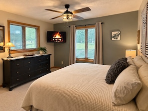 A bedroom with a bed, a dresser, a ceiling fan, a TV displaying a fireplace, and windows with curtains.