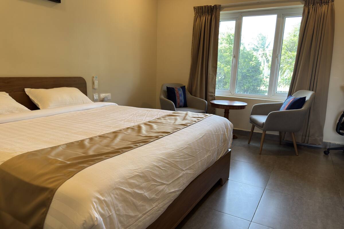 A hotel room with a large bed, a small round table, two chairs, and a window with curtains.