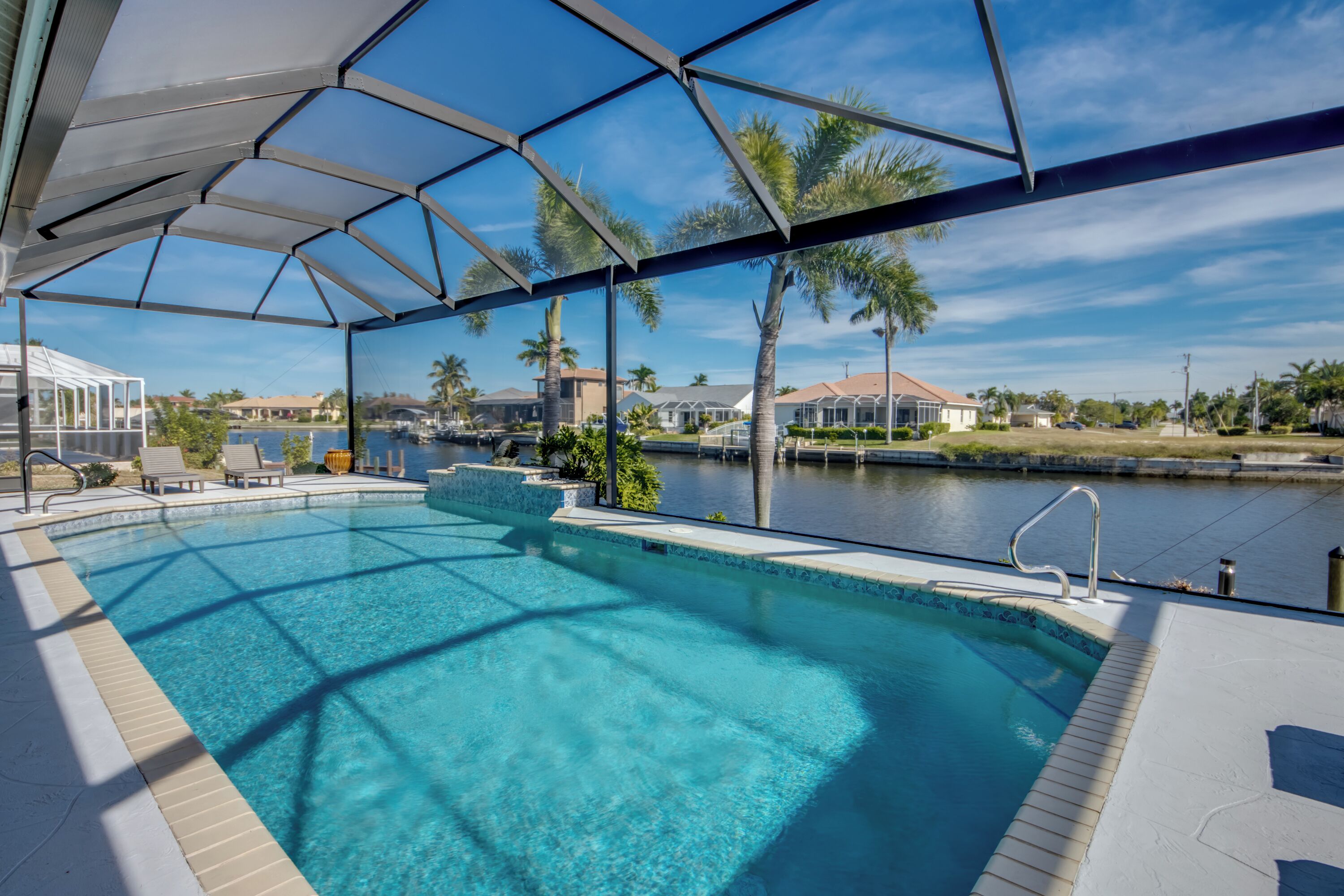 Una piscina con cerramiento de vidrio, vista al agua y palmeras al fondo.