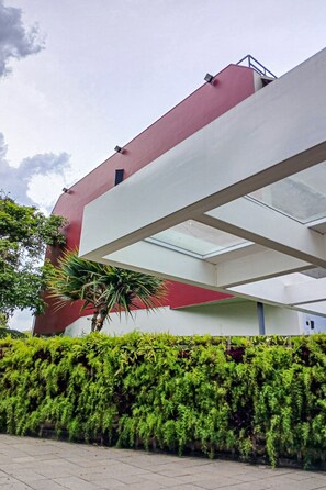A building with a red and white facade, a covered walkway, and a green hedge in the foreground. - Urbic Vila Mariana by Viva - Prédio com academia e coworking a passos da ESPM, Belas Artes e Metrô (São Paulo)