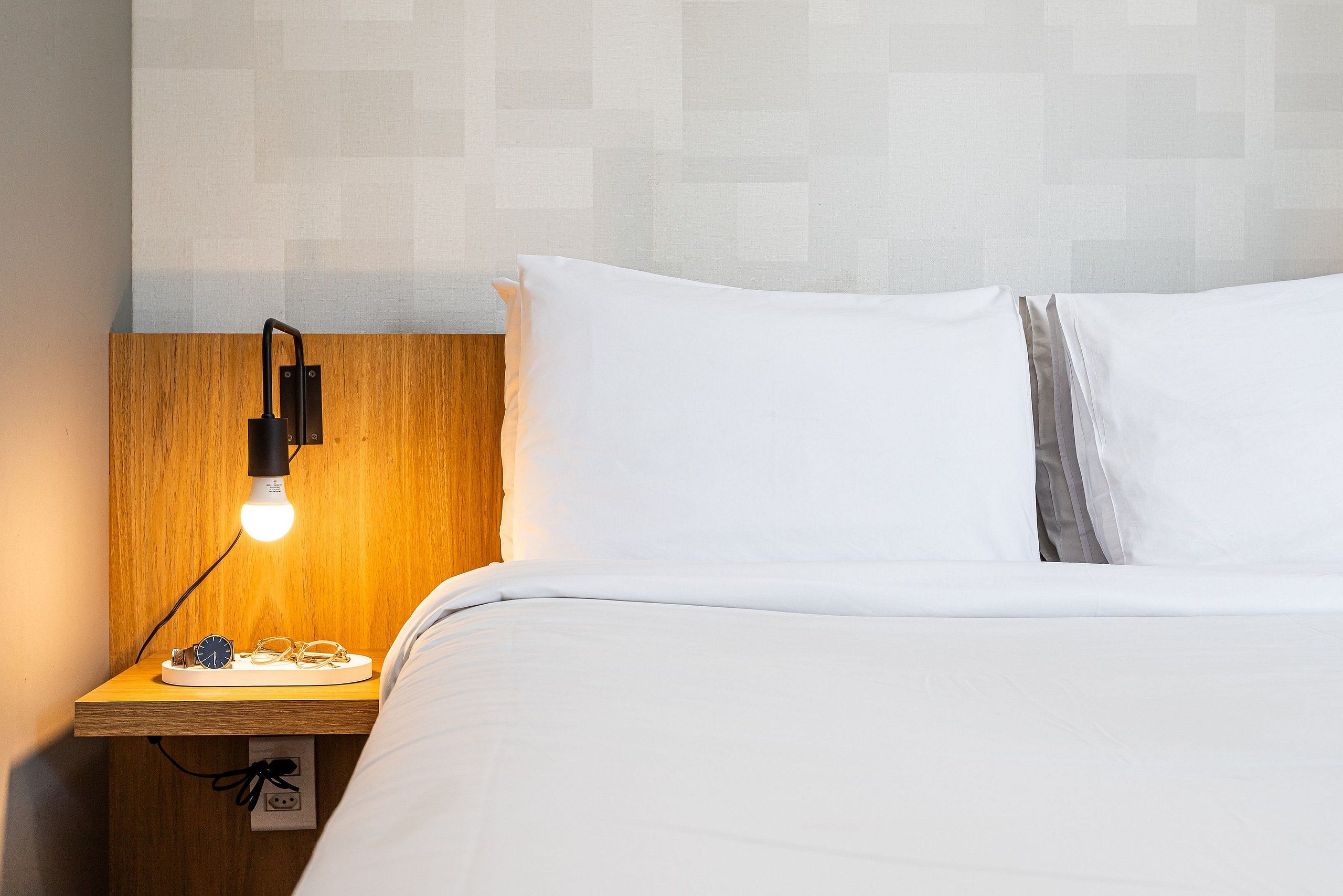 A neatly made bed with white linens, a wooden bedside table with a lamp, and a wall with a geometric pattern.