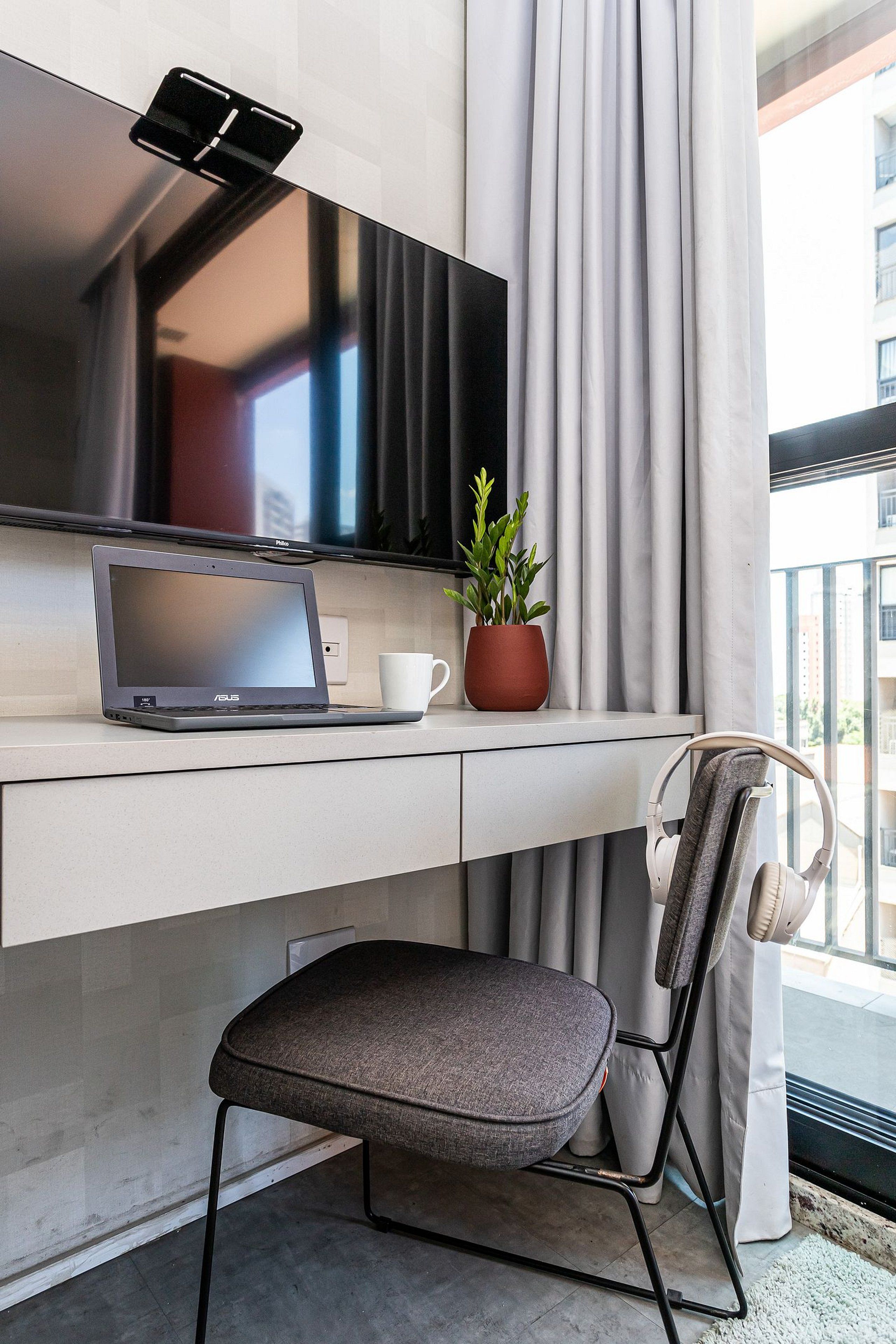 Ein modernes Hotelzimmer mit Flachbildfernseher, Laptop, Topfpflanze, weißer Tasse, Klappsessel und Kopfhörern.