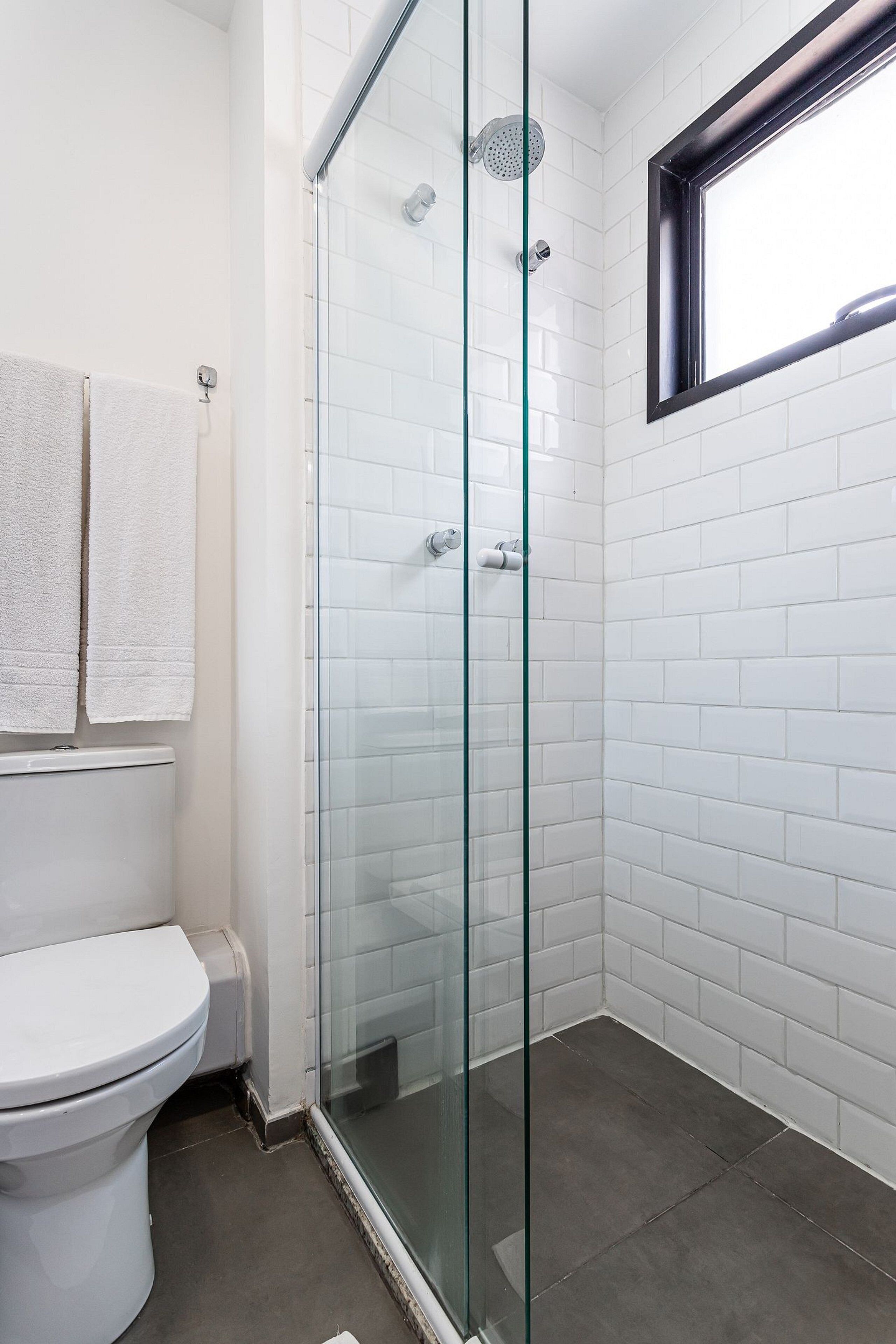 A bathroom with a glass-enclosed shower, white tiled walls, a toilet, and a window.