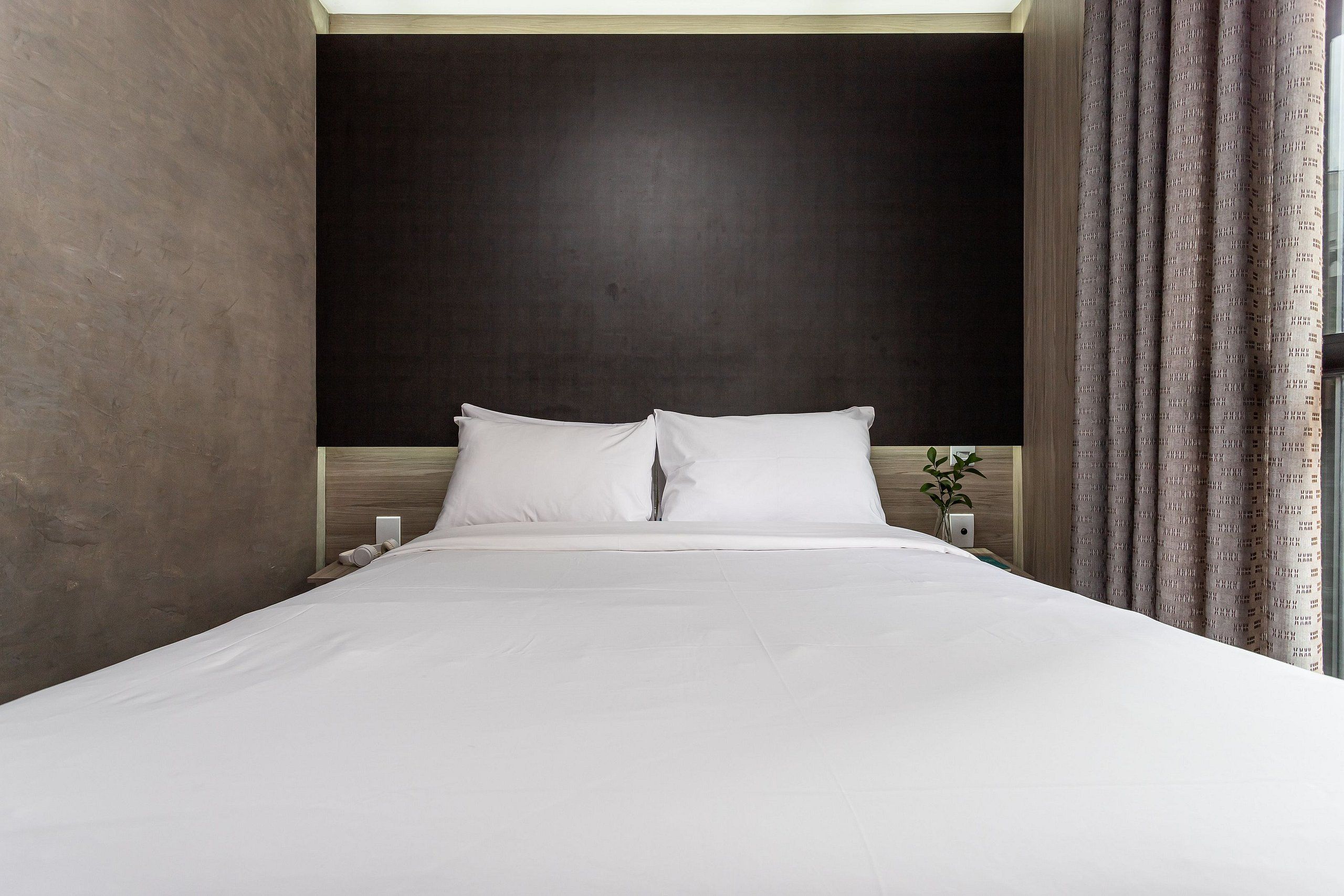 Quarto de hotel com uma cama grande, cabeceira escura e cortina no lado direito.