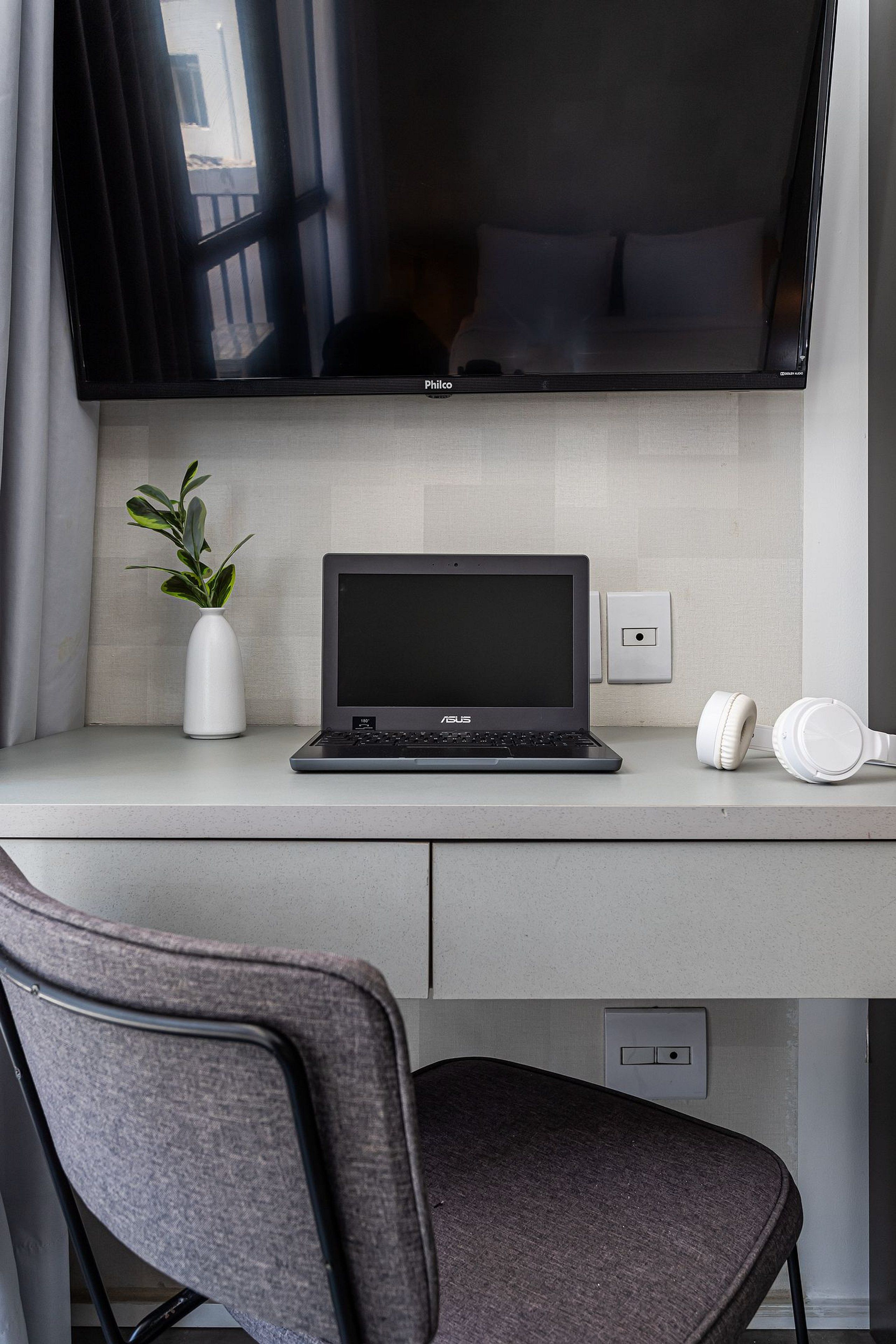 Una computadora portátil sobre un escritorio, una planta, audífonos y un televisor.