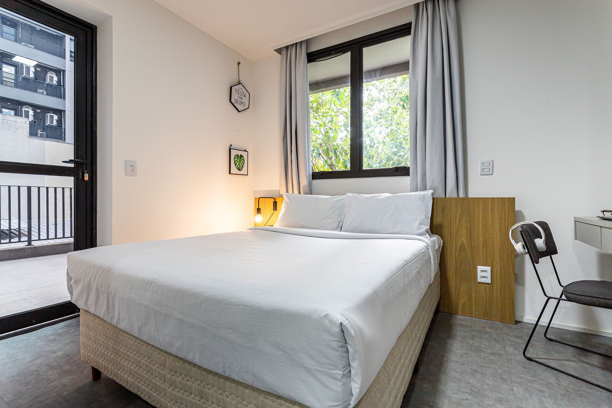 A neatly made bed with white linens, a wooden headboard, a bedside table with a lamp, a chair with headphones, and a window with curtains.