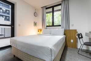 Una cama bien hecha con sábanas blancas, un cabecero de madera, una mesita de noche con lámpara, una silla con auriculares y una ventana con cortinas.