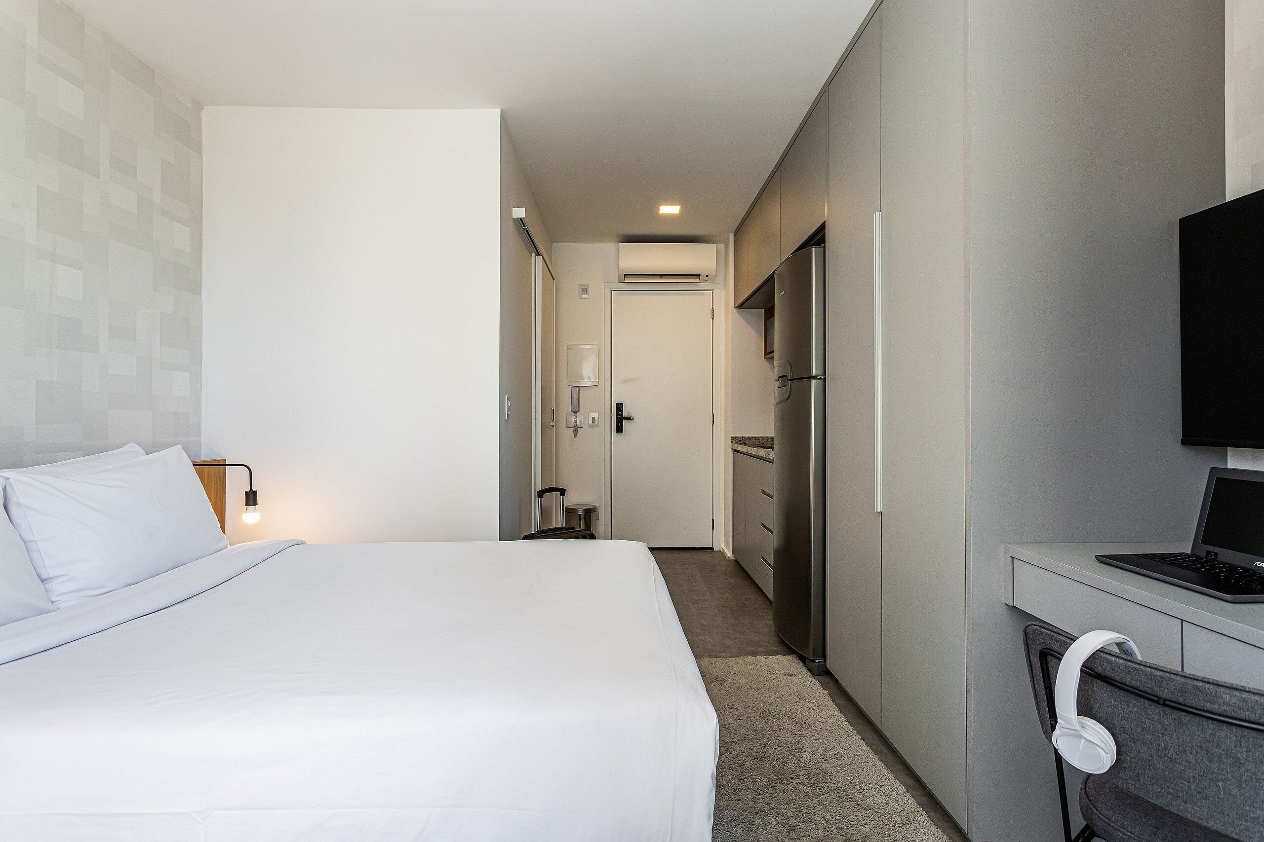 Ein Hotelzimmer mit einem Bett, einem Schreibtisch mit Laptop, einem Kühlschrank und einem an der Wand befestigten Fernseher.