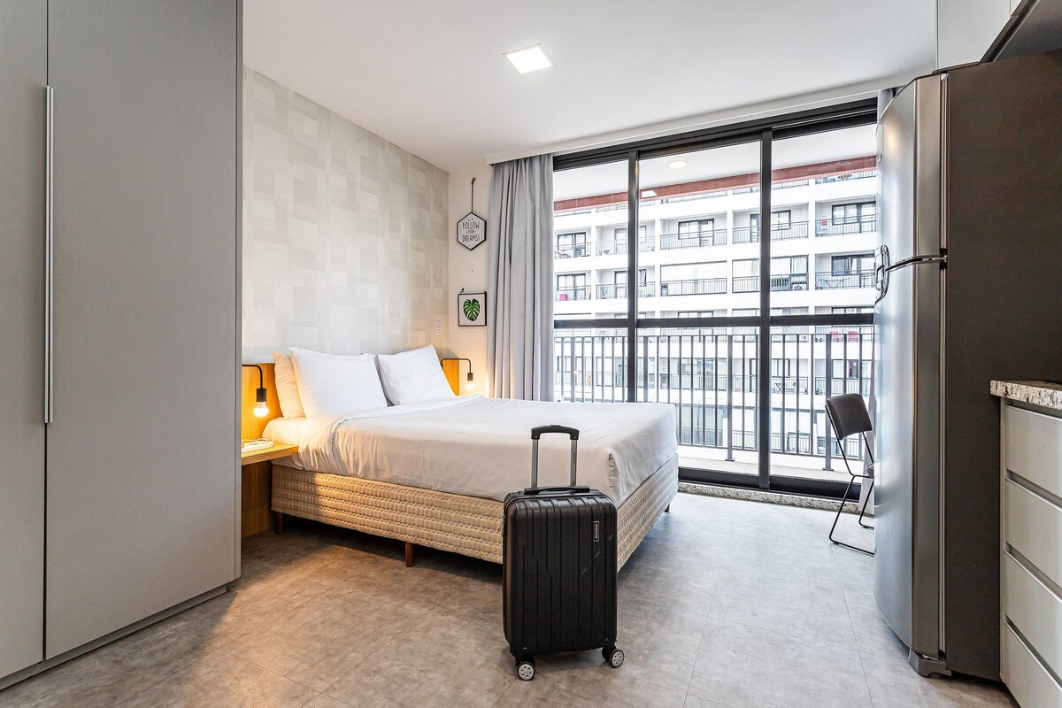 Un cuarto de hotel con una cama, una maleta, un escritorio con una lámpara, una silla y un ventanal con vistas a un edificio.