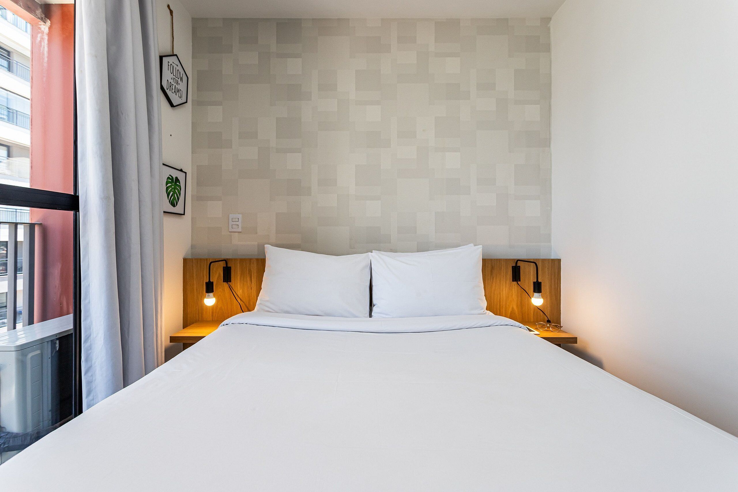 Ein ordentlich gemachtes Bett mit weißen Leinen, zwei Nachttischlampen, einem Holz-Kopfteil und einer Wand mit geometrischem Muster.