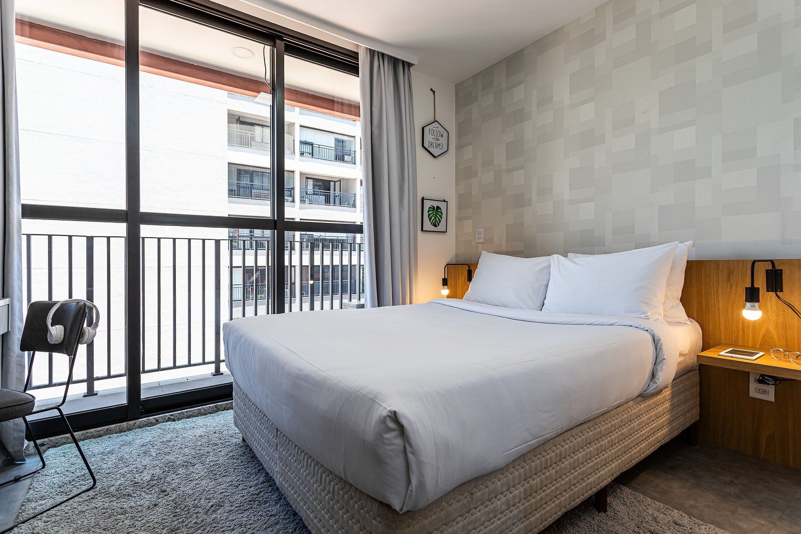 Ein Hotelzimmer mit einem großen Bett, einem Schreibtisch mit Kopfhörern sowie Blick auf einen Balkon und einer Wand mit geometrischem Muster.