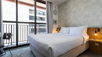 Ein Hotelzimmer mit einem großen Bett, einem Schreibtisch mit Kopfhörern sowie Blick auf einen Balkon und einer Wand mit geometrischem Muster.