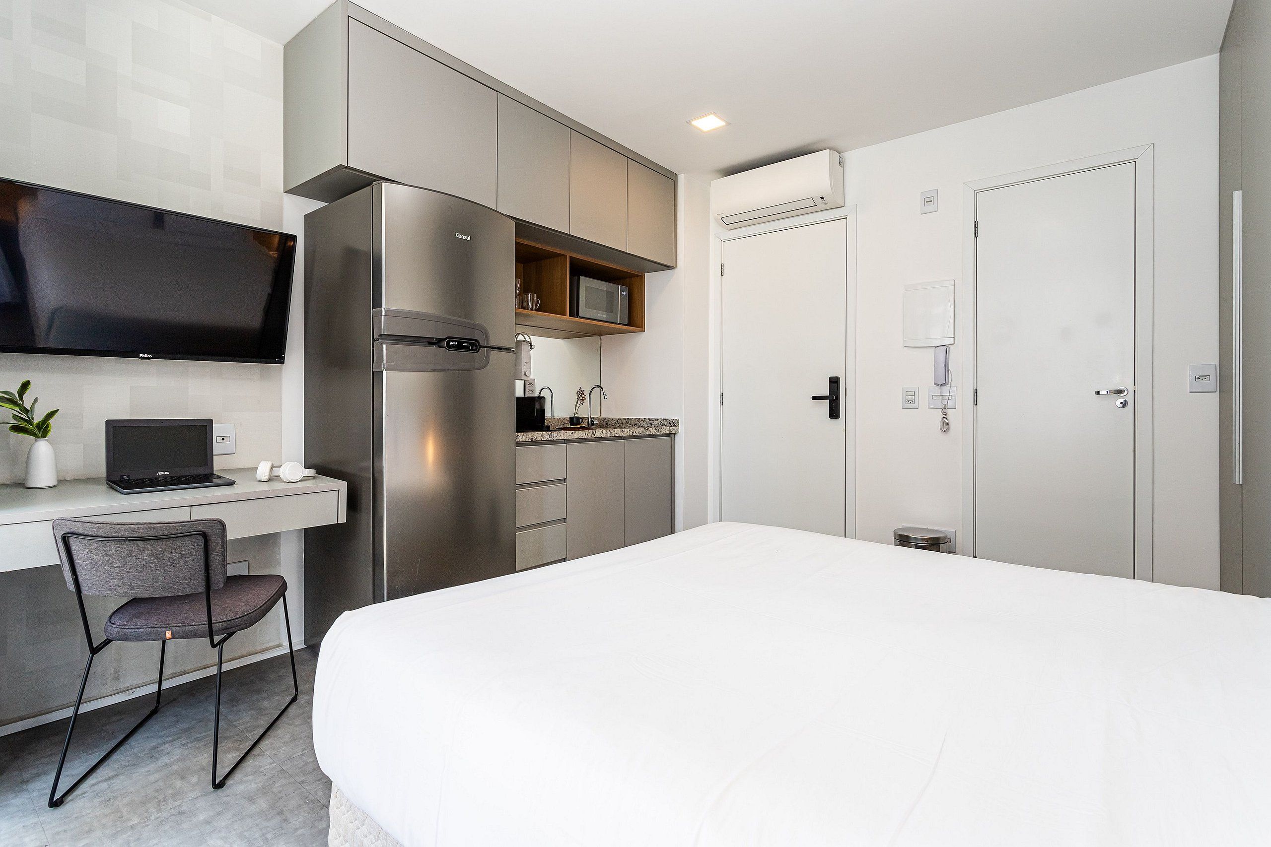 A modern hotel room with a bed, a desk with a laptop, a chair, a refrigerator, a microwave, and a television.
