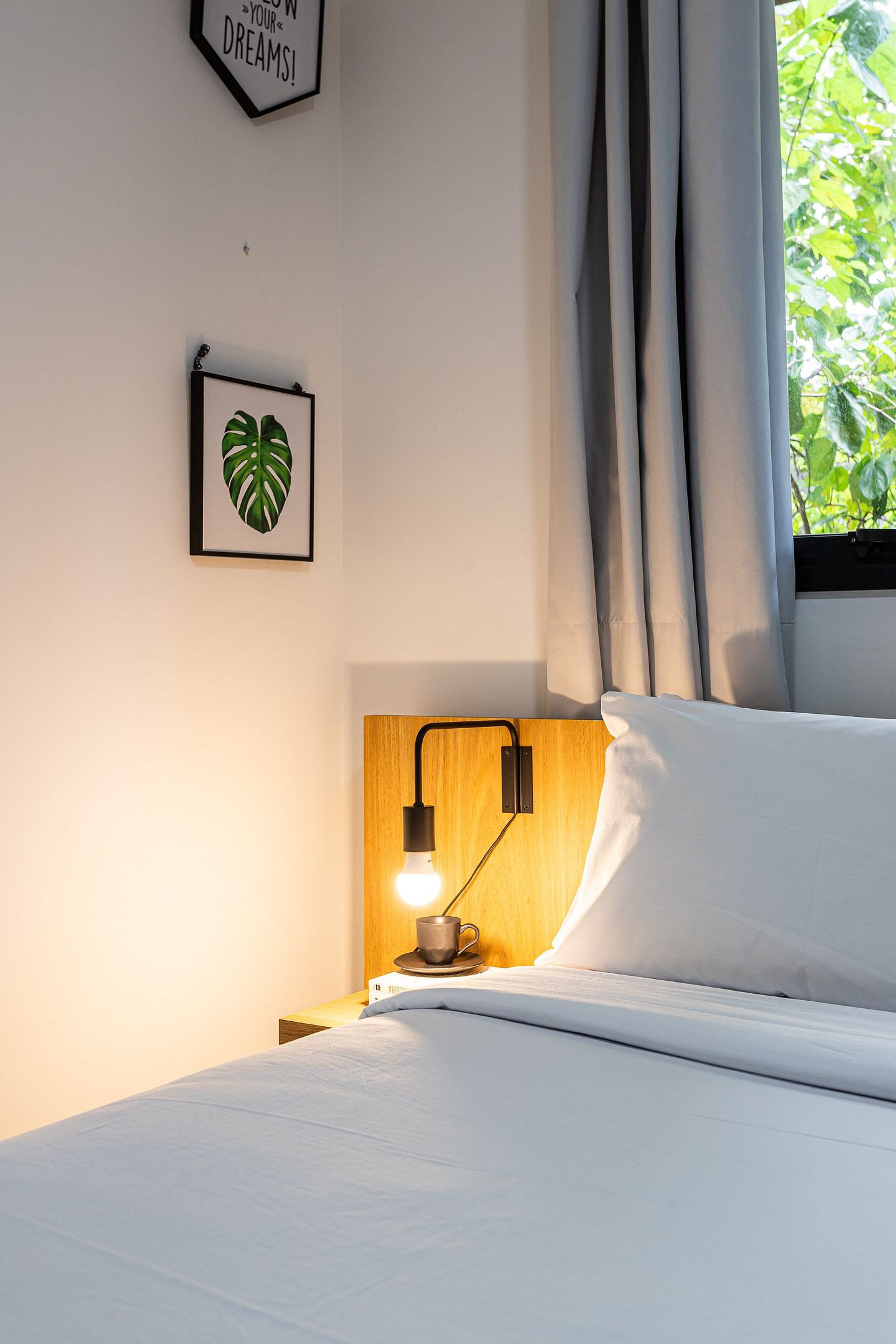 Un lit bien fait, avec un oreiller blanc et une tête de lit, une table de chevet avec une lampe, une œuvre d’art représentant une feuille encadrée, et une citation inspirante sur le mur.