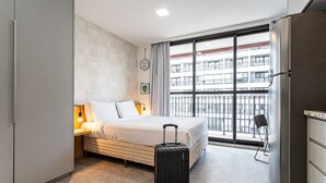 A hotel room with a bed, a suitcase, a desk with a lamp, a chair, and a large window with a view of a building. - Urbic Vila Mariana by Viva - Prédio com academia e coworking a passos da ESPM, Belas Artes e Metrô (São Paulo)