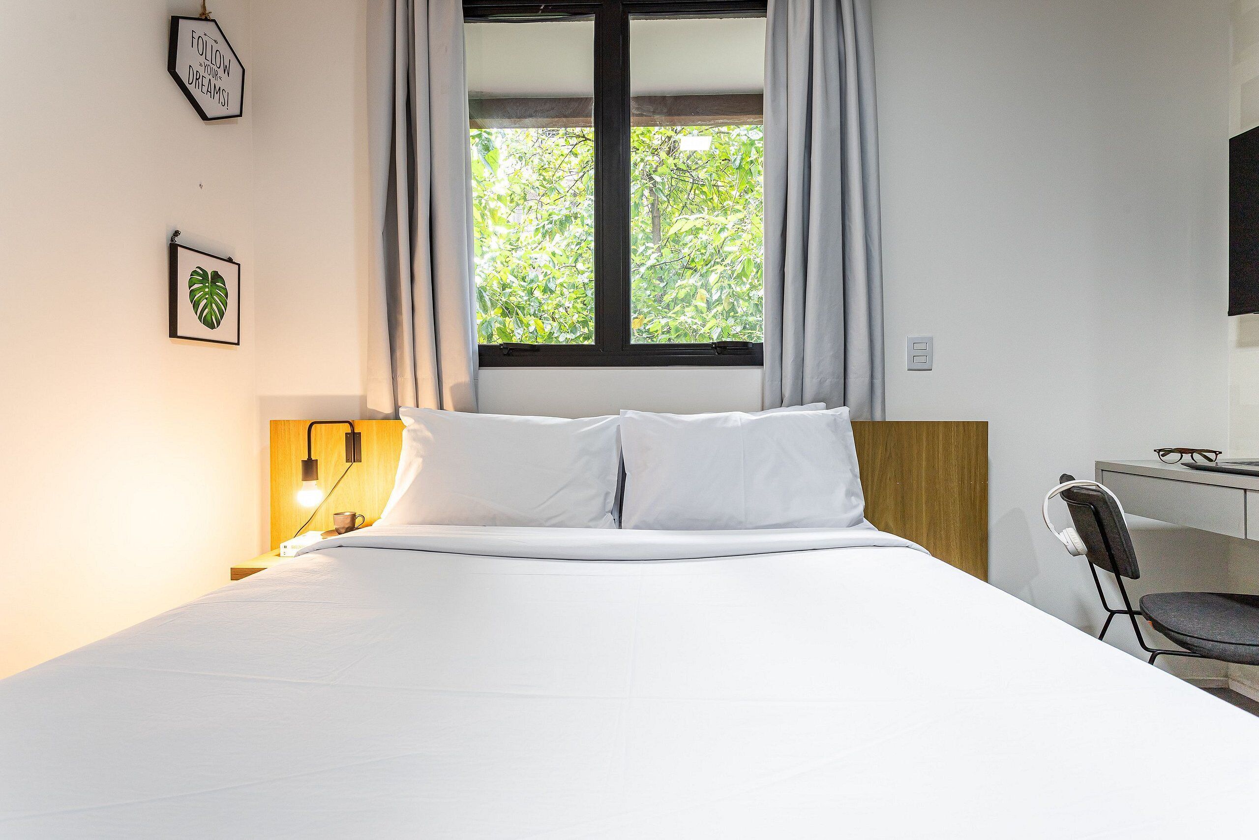 A neatly made bed with white linens, a desk with a chair, a wall-mounted TV, and a window with curtains.