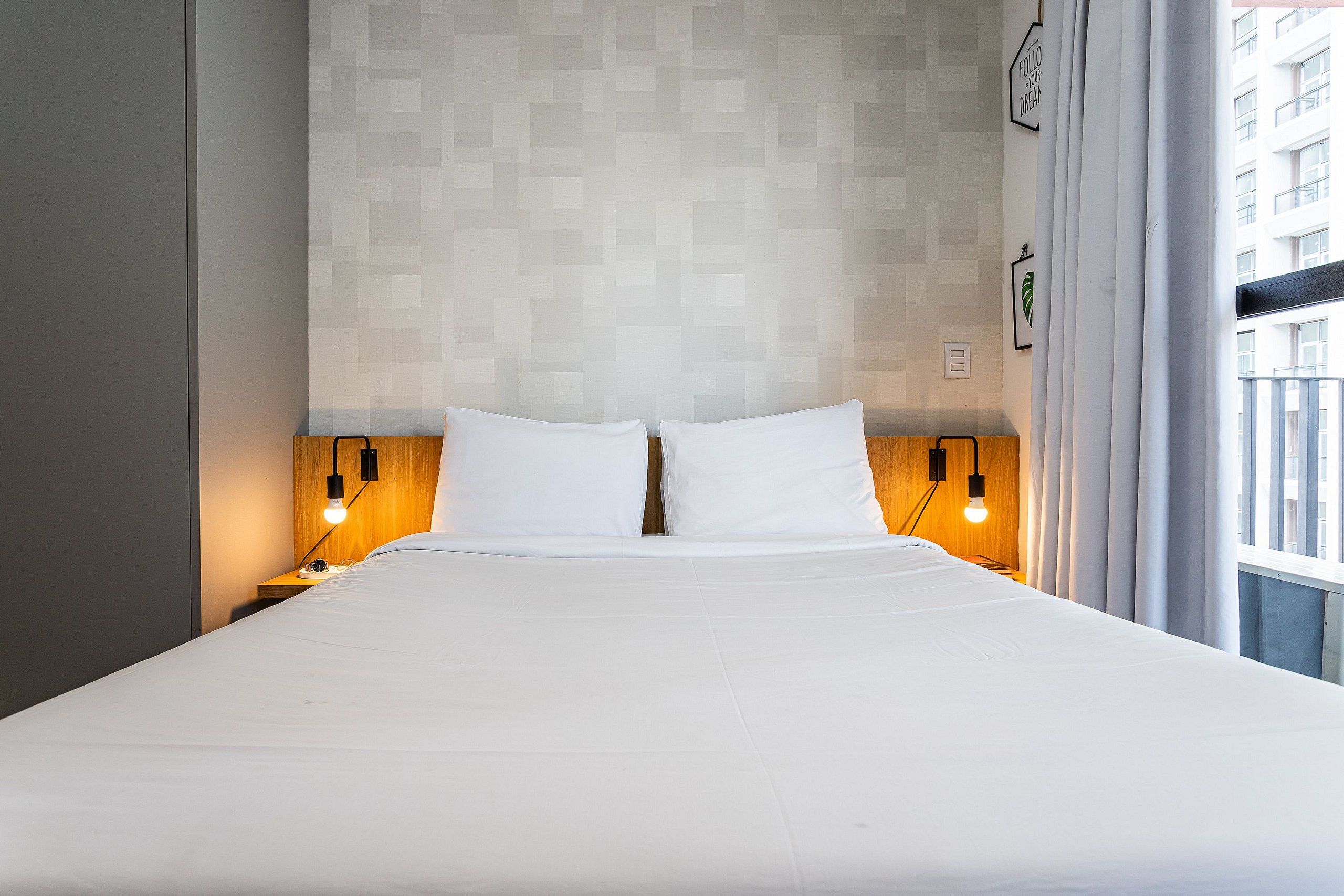 Una habitación de hotel con una cama grande, dos mesitas de noche con lámparas, una pared con diseño geométrico y una ventana con cortinas.