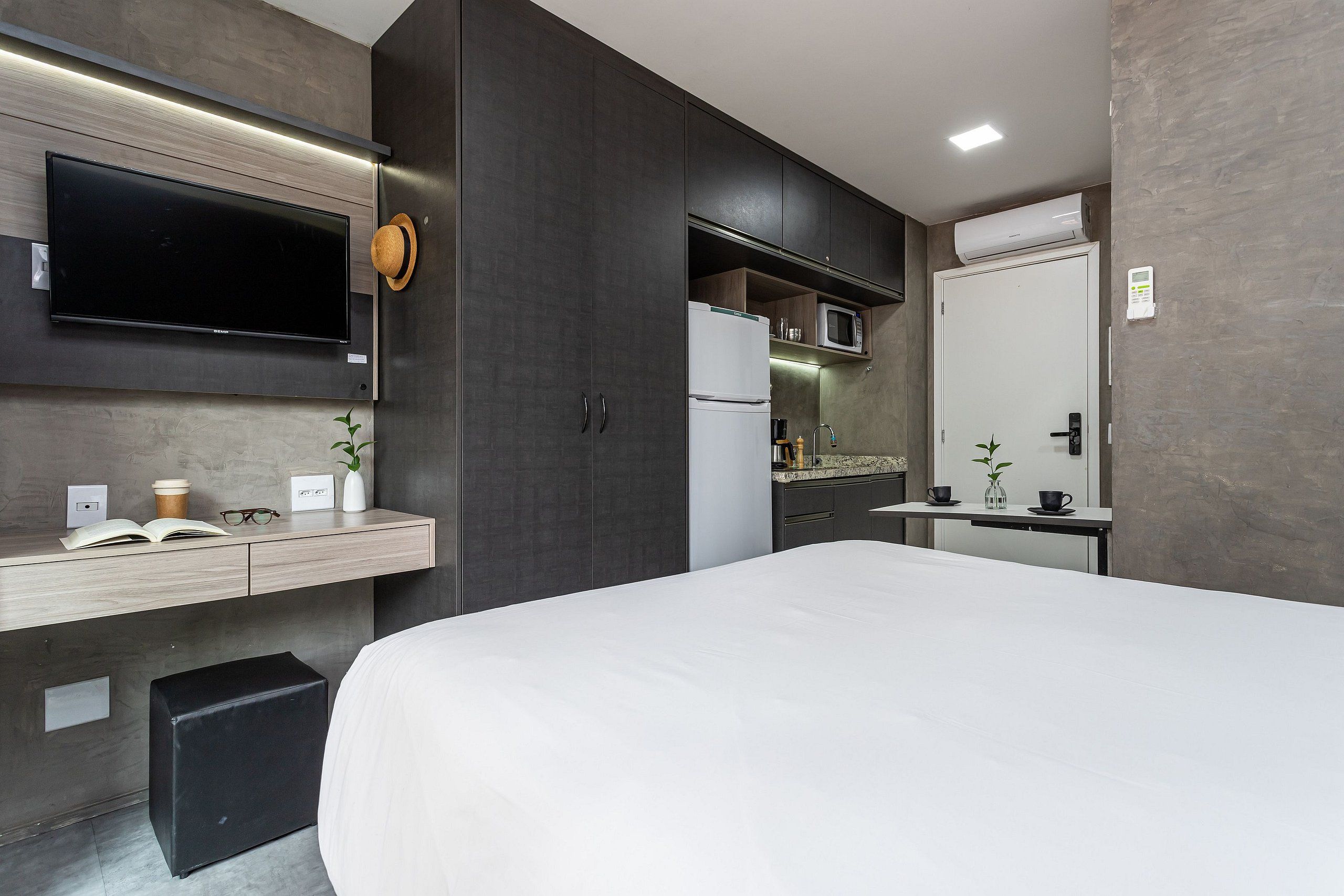 A modern hotel room with a large bed, a flat-screen TV mounted on the wall, a small kitchen area with a microwave, and a separate bathroom with a toilet and sink.