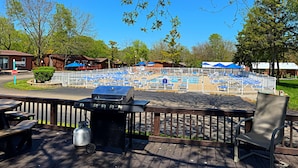 Zona de piscina amb ombrel·les blaves, una barbacoa i taules de picnic sobre un pont de fusta.