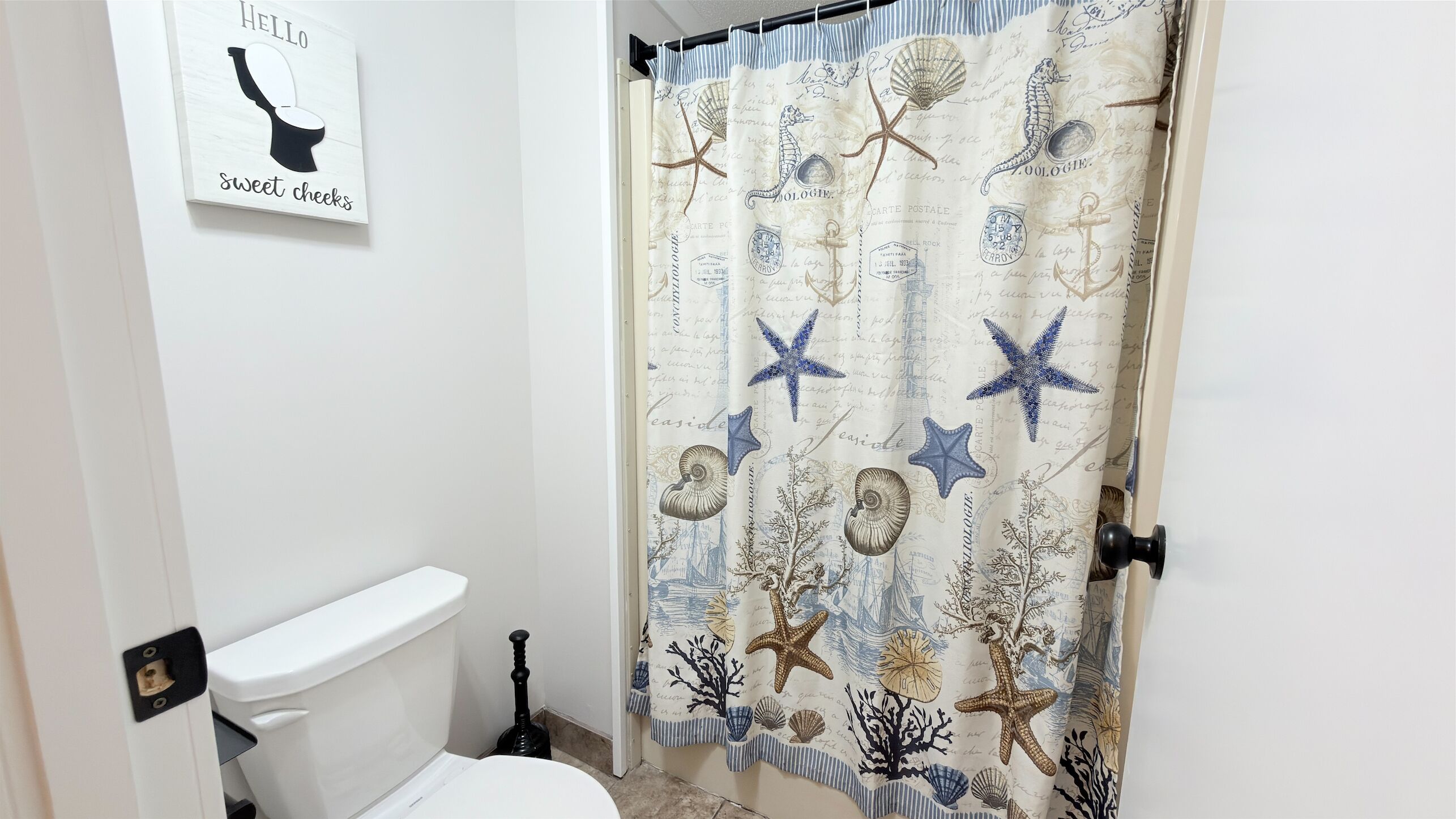 A bathroom with a toilet, a plunger, and a shower curtain with nautical designs.