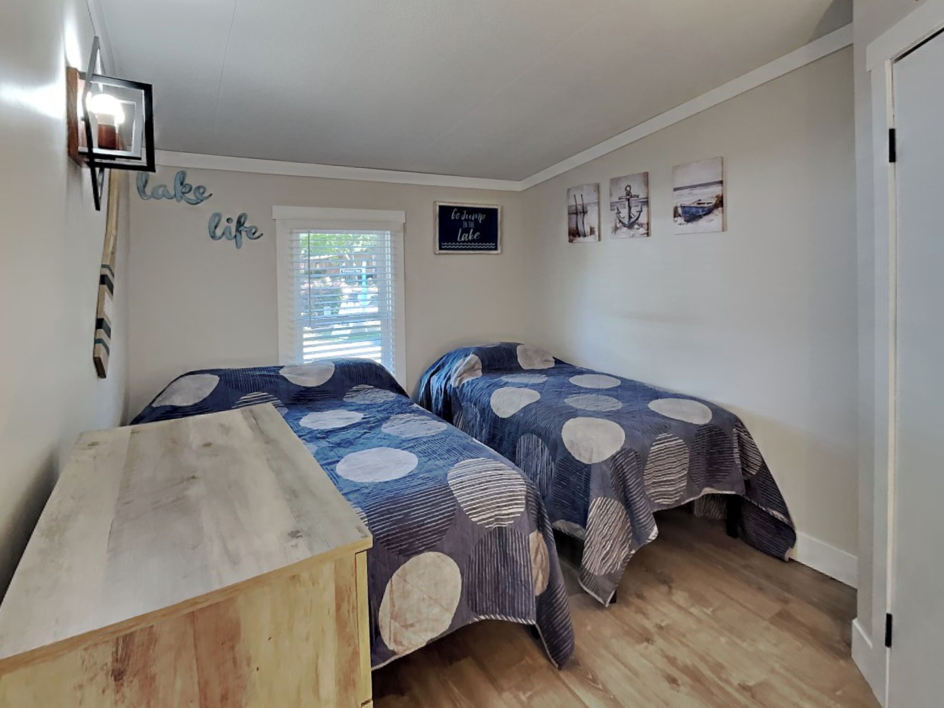 A bedroom with two beds, a wooden dresser, a window with blinds, and nautical-themed wall art.