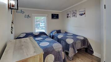 A bedroom with two beds, a wooden dresser, a window with blinds, and nautical-themed wall art.