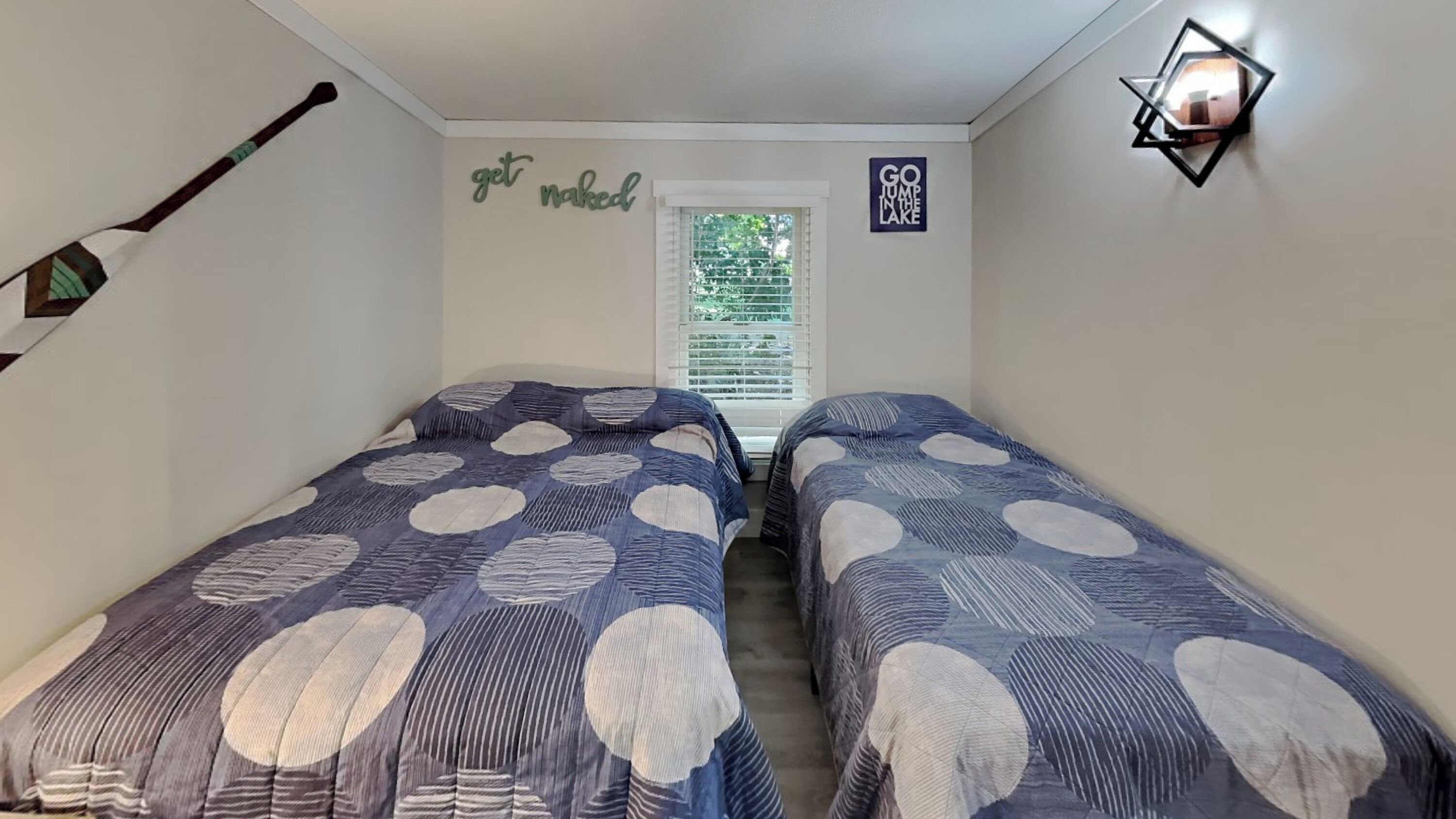 A small, narrow room with two beds, a window with blinds, and a wall with motivational signs.