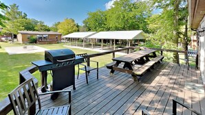 En træterrasse med picnicborde og stole, grill og overdækkede picnicområder i baggrunden.