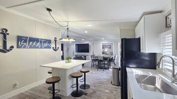A modern kitchen with a central island, bar stools, a stainless steel sink, and a dining area with a TV.